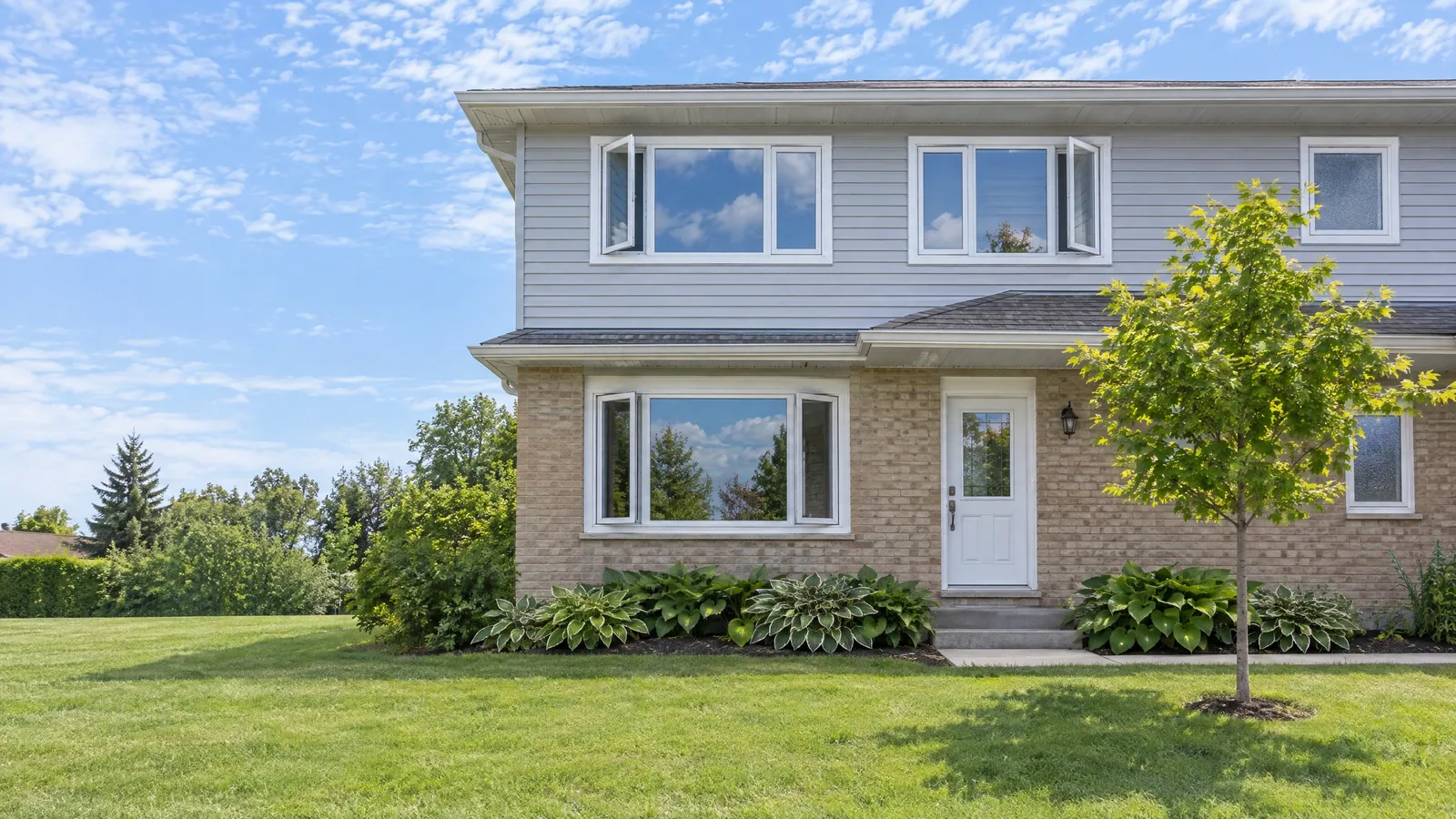 Two-storey house with buff brick below and grey siding above, fitted with white vinyl casement windows, several cranked open on a summer day
