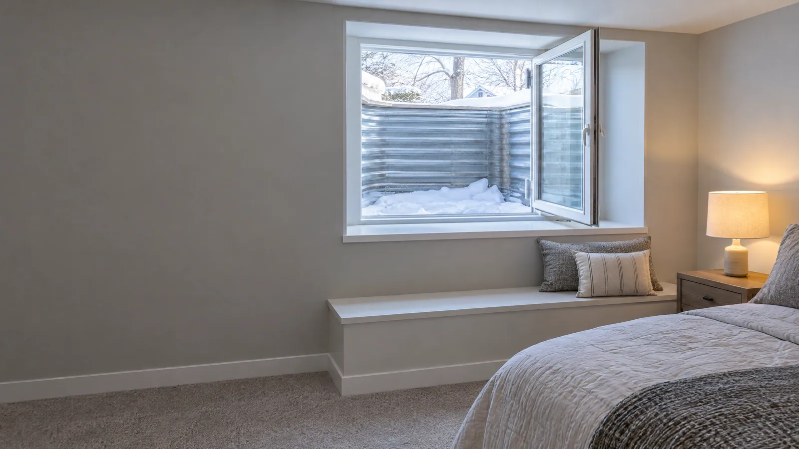 Finished basement bedroom with a wide casement window swung open onto a corrugated steel window well full of snow, above a built-in bench