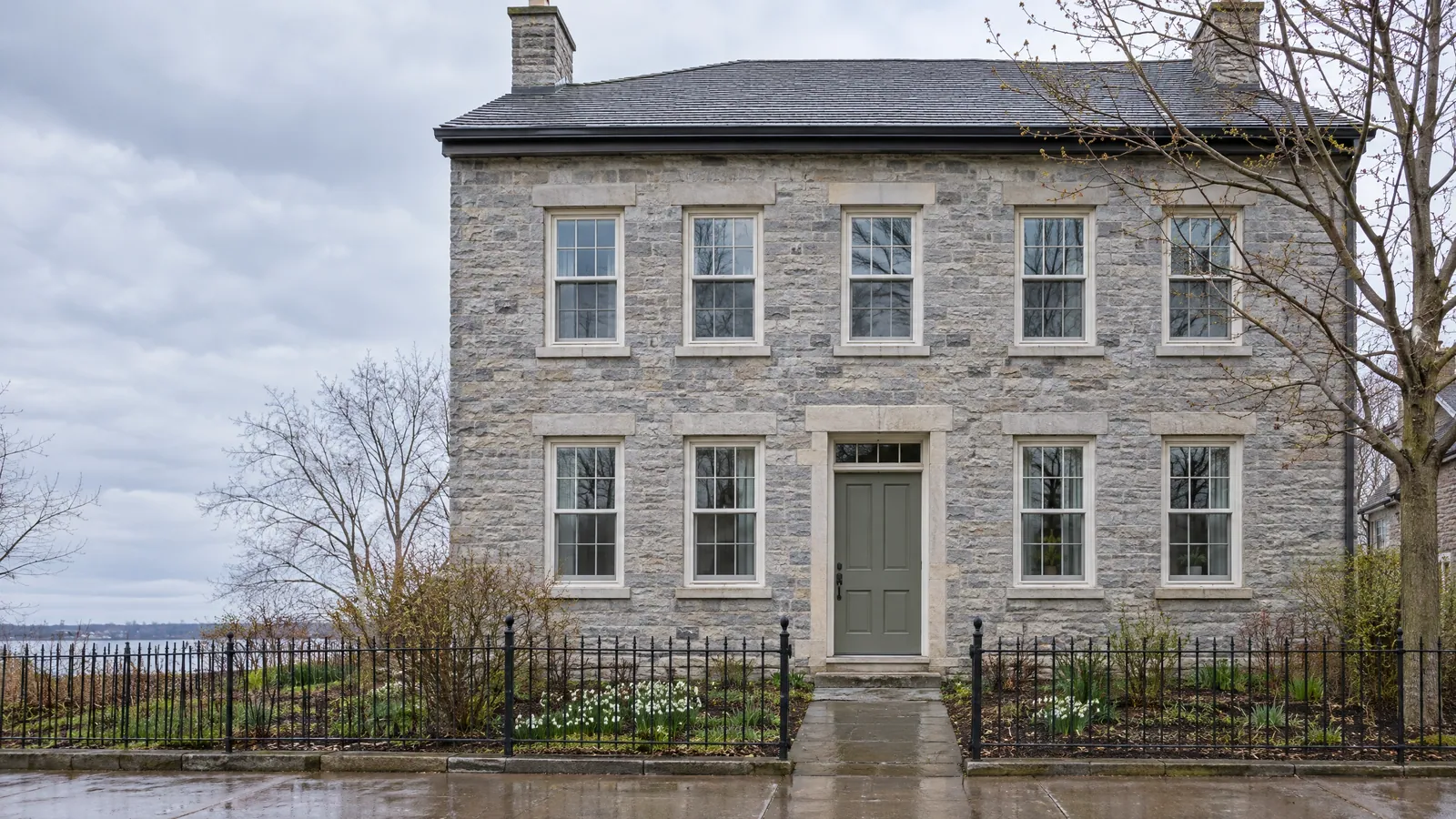 Symmetrical two-storey grey limestone house with dressed stone lintels and sills, deep-set six-over-six wood sash windows, a transom over the front door and an iron fence, with the lake behind on an overcast spring day