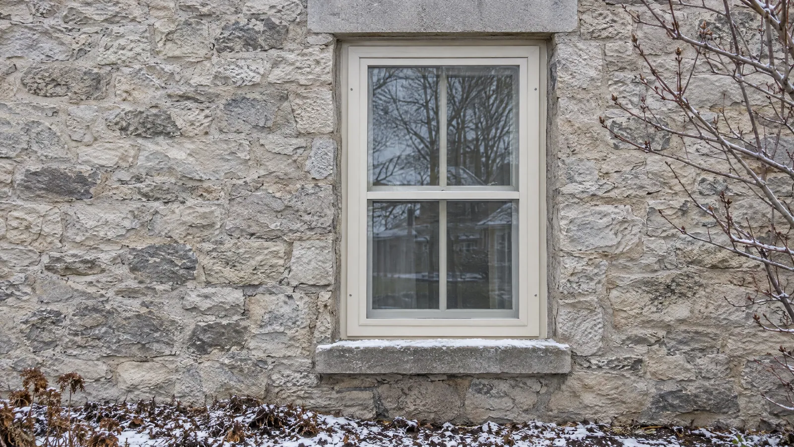 Cream-painted storm window over a two-over-two sash in a rough-cut limestone wall, with a dressed stone lintel and a dusting of snow on the stone sill