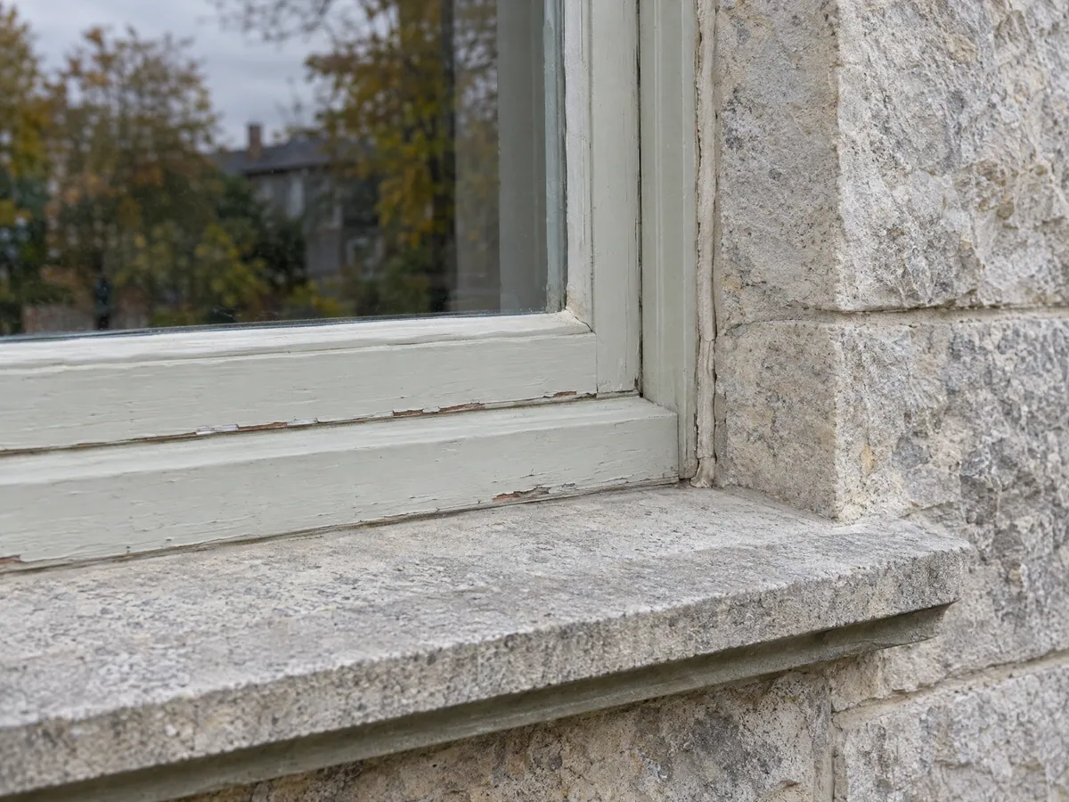 Close-up of a pale green painted wood window corner, with paint flaking along the bottom rail, set against a rough-faced limestone jamb above a smooth dressed stone sill